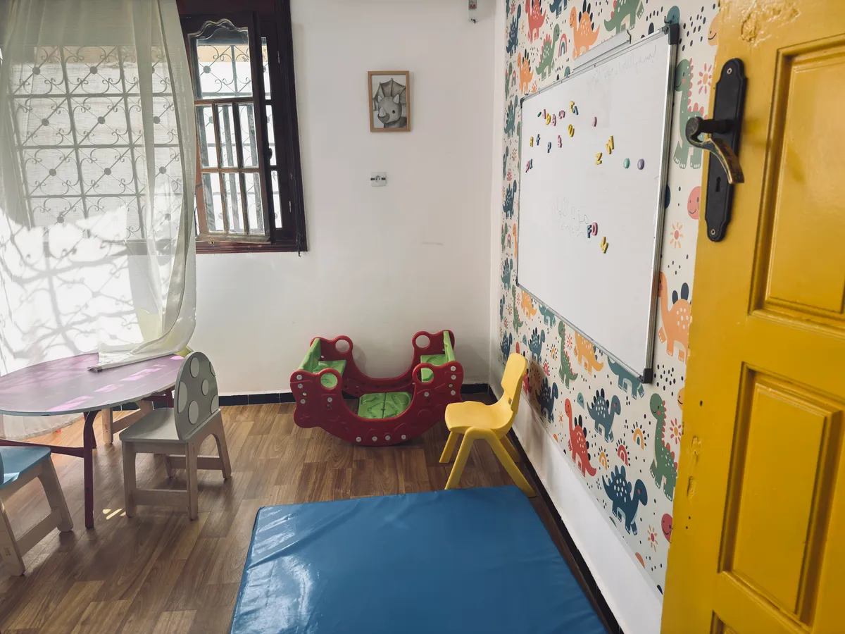 Room with a large blue mat, a rocking toy, a small table and a whiteboard.