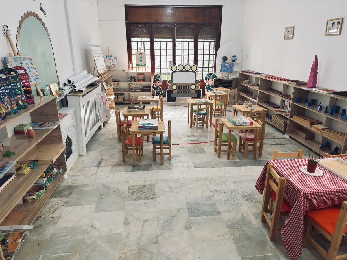 Large, bright Montessori room with low shelves and small wooden tables and chairs.