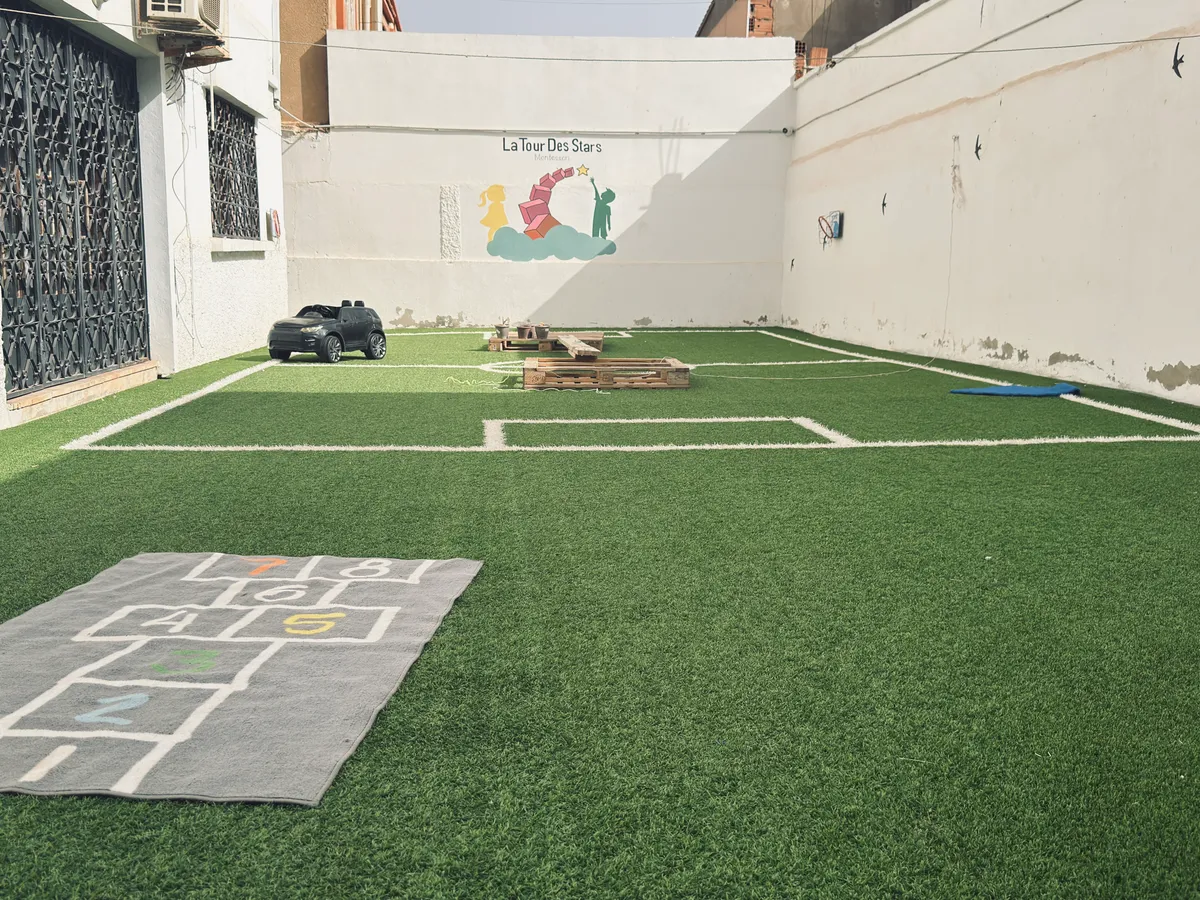 Outdoor yard with artificial grass, a hopscotch mat, a toy car and the nursery mural in the background.