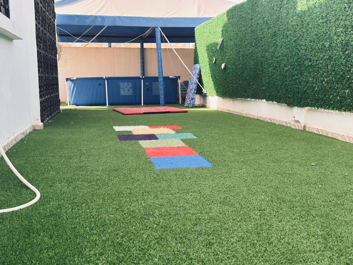 Terrace with artificial grass, colourful tiles, a green hedge and a covered area at the back.